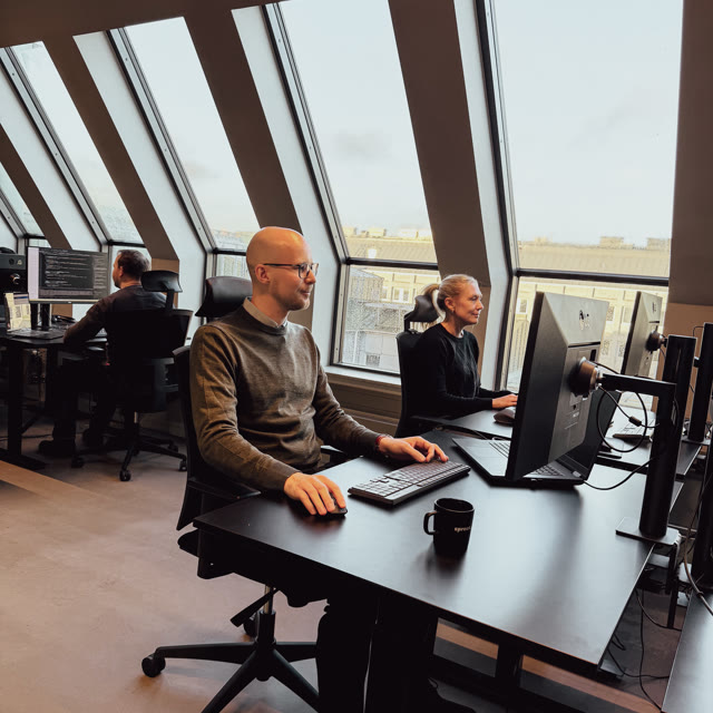 Personer arbetar vid skrivbord med datorer i ett modernt kontorslandskap med stora fönster och dagsljus.