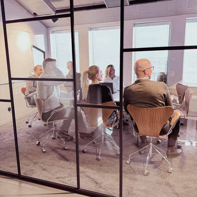 Sex personer sitter vid ett bord och diskuterar arbete vid sina bärbara datorer i ett mötesrum på ett kontor.