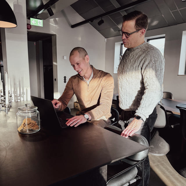 Två personer står vid ett bord och arbetar tillsammans vid en bärbar dator i en ljus kontorsmiljö.